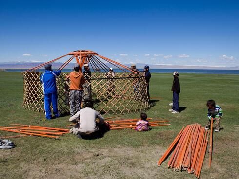 A group of people build a yurt with lattice wood walls on a grassy landscape