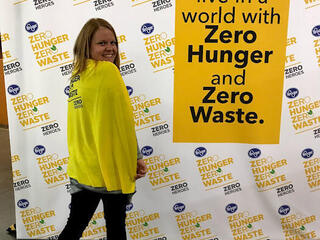 A woman stands in front of a "Zero Hunger | Zero Waste" sign