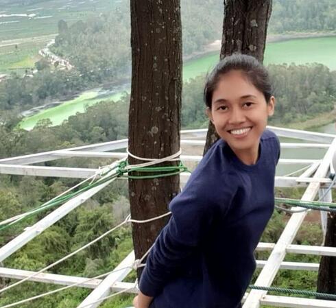 Nety Riana Sari stands beside a tree overlooking a forest and smiles at the camera