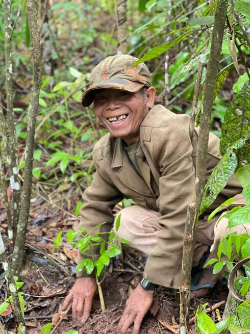 Older man in a hat crouching and smiling while planting a tree in Laos