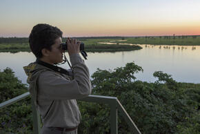Carolina Alvarez looks out across a river with binoculars at sunset