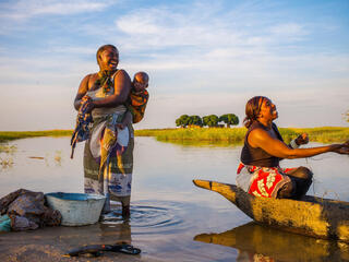 Two women and a baby on a river bank, one washing clothes and the other casting a net while all smile
