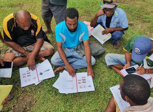 Men sit in a semicircle on grass looking at notebooks with fish images in them