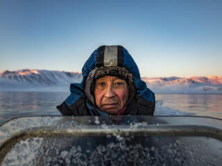 A man wearing a thick blue parka looks into the camera. He is on a boat at sunrise. Behind him is snow covered land.